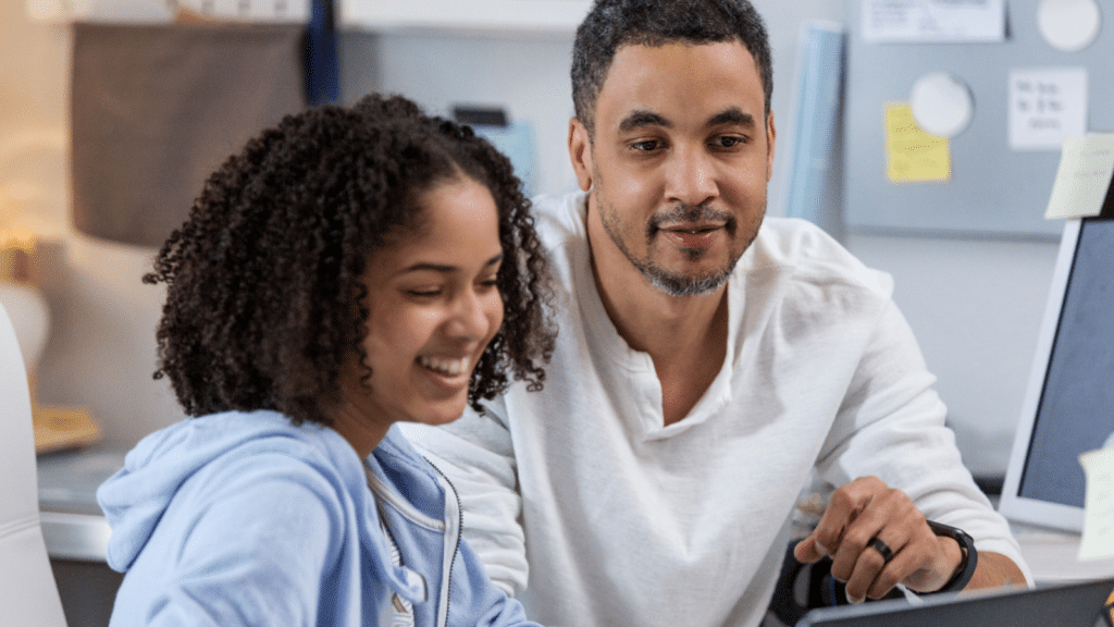 A man and a young girl smile while looking at a laptop screen, engaged in STEM education, in a modern office setting.