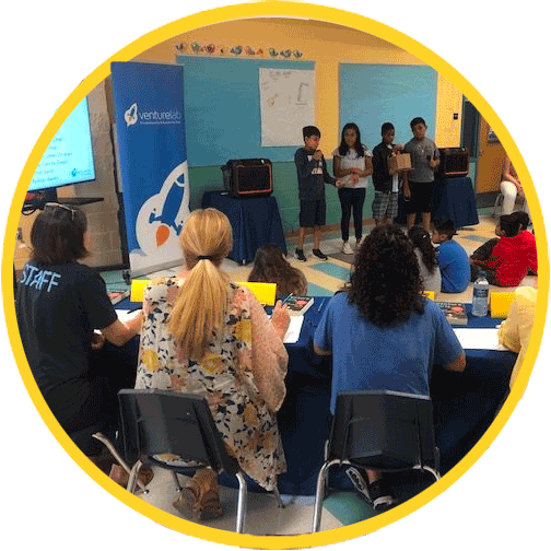 Group of lower elementary children use VentureLab Curriculum & Programs to present a project in a classroom with audience watching and taking notes, educational banners in the background.